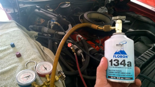 Technician inspecting car AC system with R-134a refrigerant cylinder nearby.