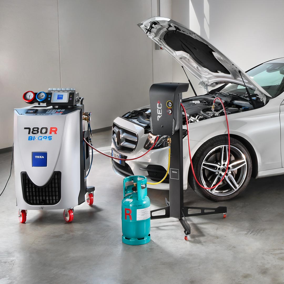 Automotive A/C service setup showing a Mercedes-Benz with its hood open connected to TEXA 780R Bi-Gas recovery and charging station, refrigerant recovery unit, and a turquoise R-type refrigerant cylinder in a clean workshop.