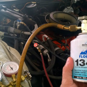 Technician inspecting car AC system with R-134a refrigerant cylinder nearby.